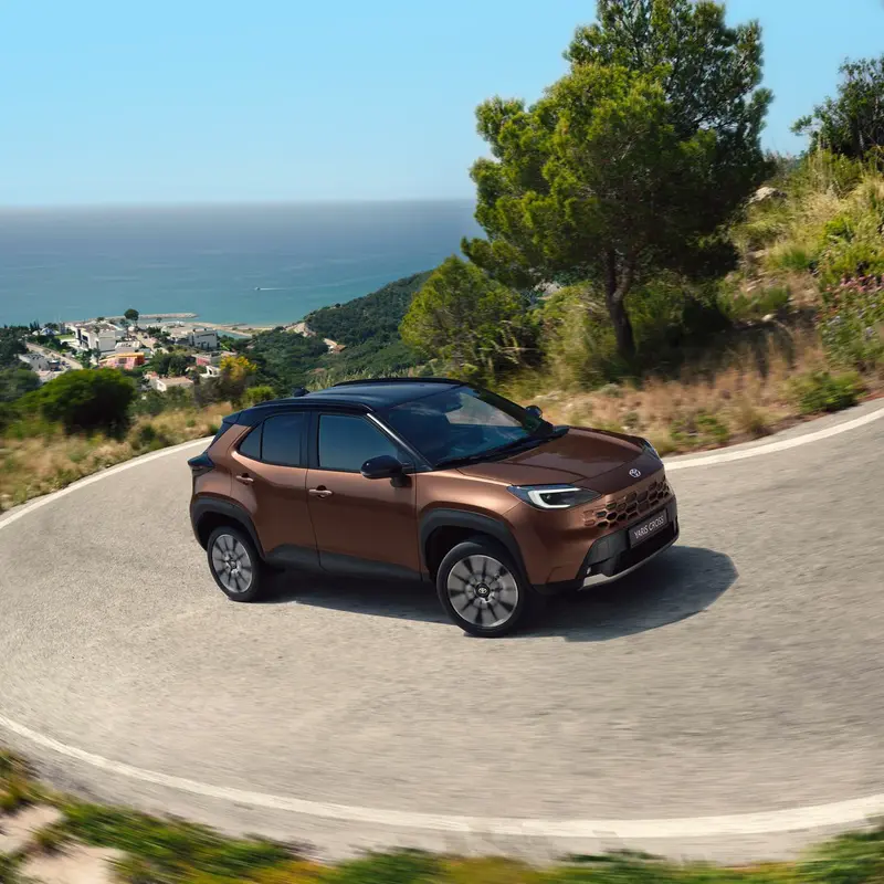 Bruine Toyota Yaris Cross in een bocht op een kustweg met zicht op zee